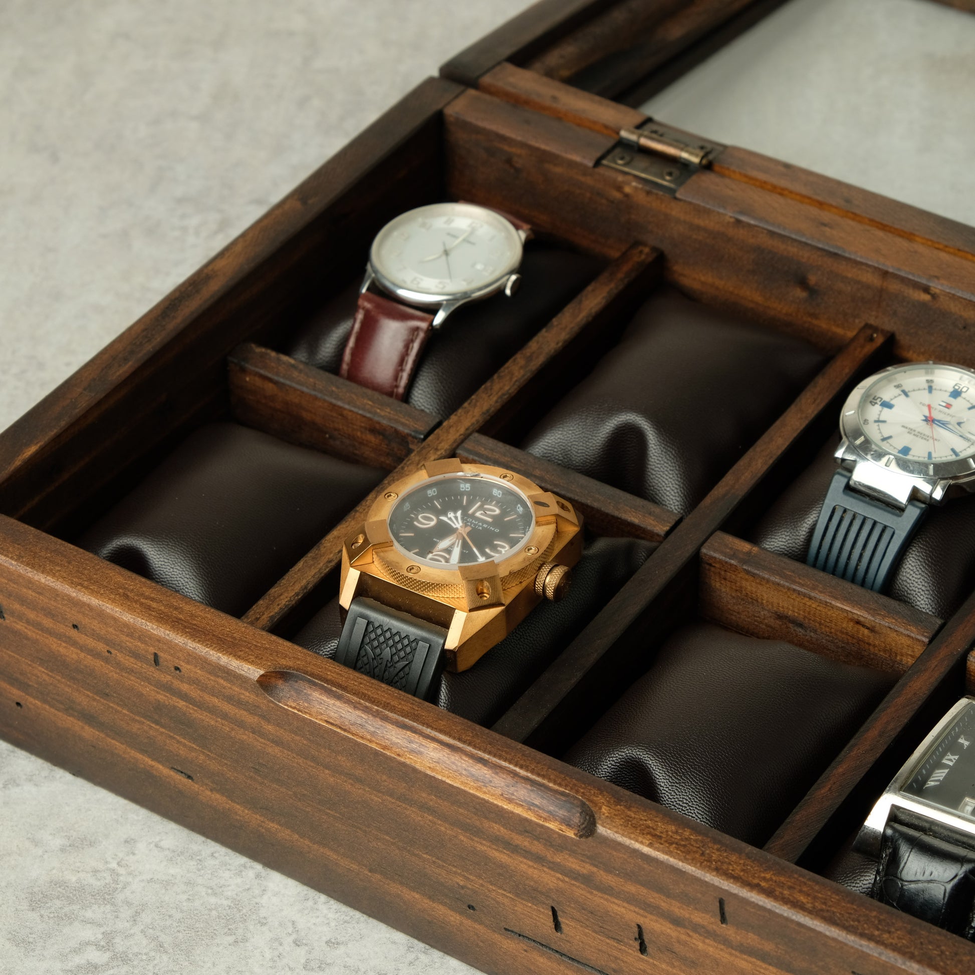 A wooden watch display box with multiple cushioned compartments holding several wristwatches, photographed on a light gray surface.