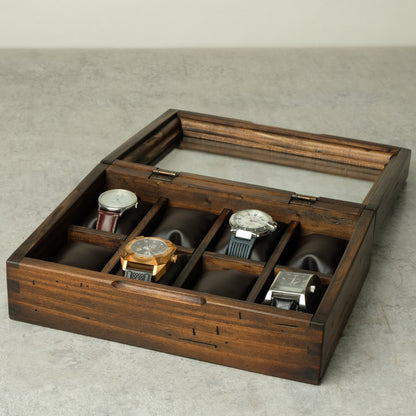 A dark brown wooden watch box with a glass lid, shown open and containing several watches on dark cushion inserts, set against a neutral gray background.