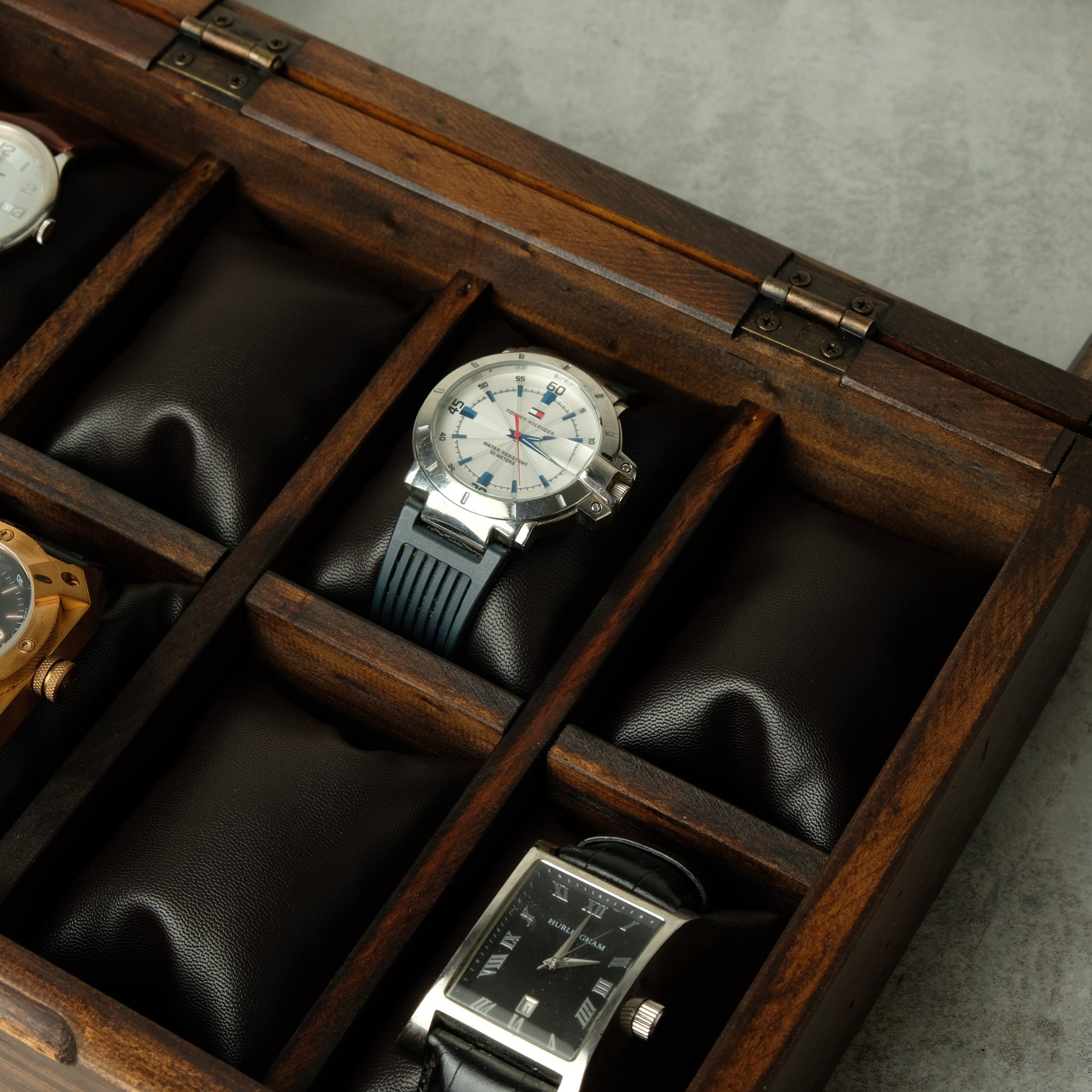 A wooden watch display box with six cushioned compartments, shown open and containing several wristwatches. The box has a dark brown wood finish with metal hinges and is photographed on a light gray surface.
