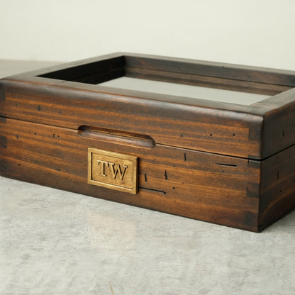 A dark brown wooden display box with a glass top, featuring a brass monogrammed plate