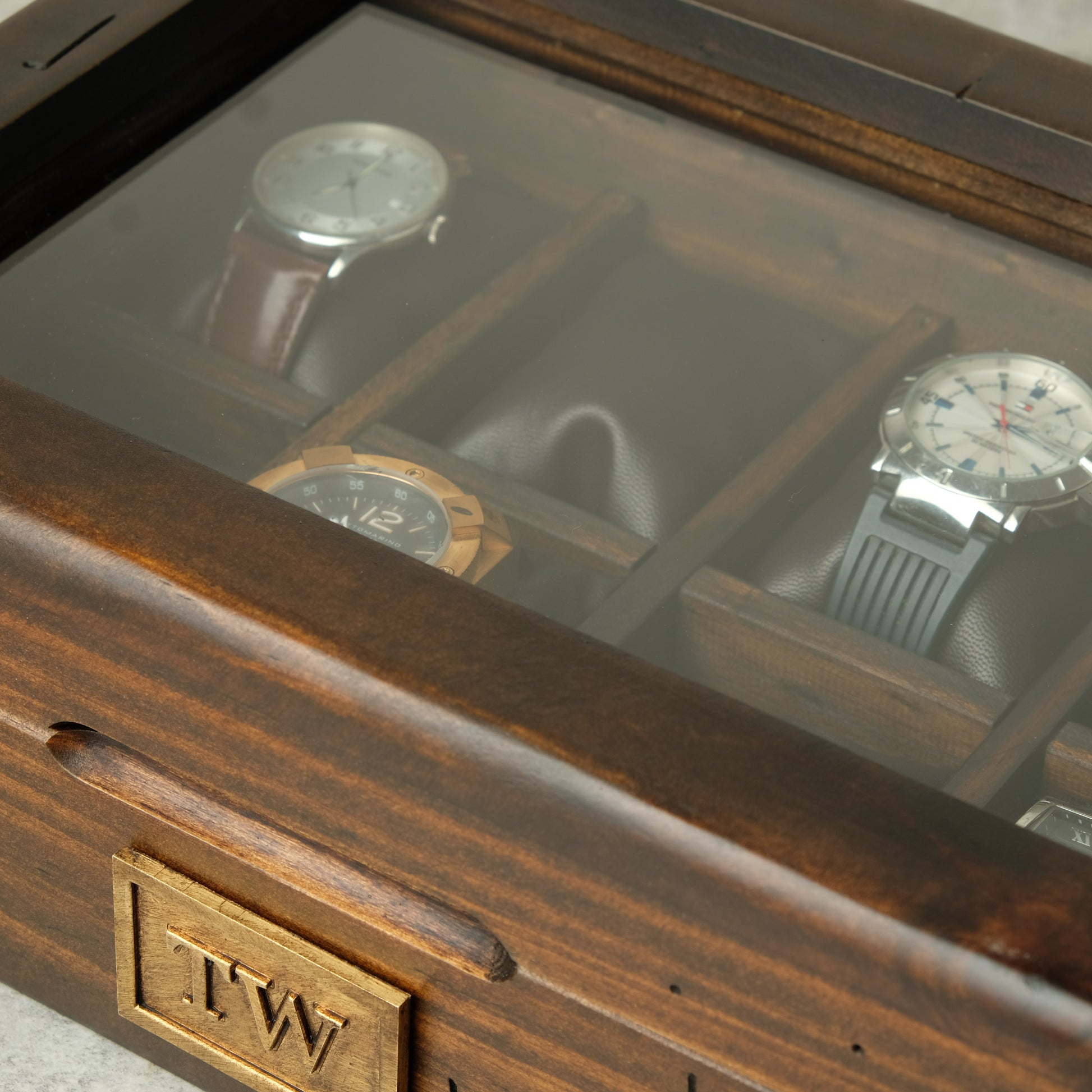 Close-up of a wooden watch display case with a glass top, showing several watches stored inside. The front corner has an engraved wooden plaque with the initials 'T.
