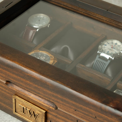 Close-up of a wooden watch display case with a glass top, showing several watches stored inside. The front corner has an engraved wooden plaque with the initials 'T.