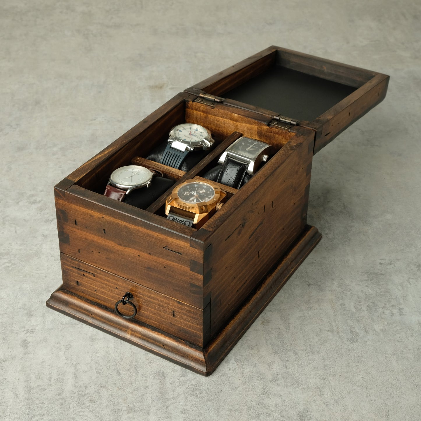 A dark brown wooden watch box with an open lid, containing four watches on black cushions, set against a neutral gray concrete background.