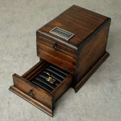 A dark brown wooden jewelry box with one drawer pulled open, showing a black ring roll holding a gold ring. The box has a metal plaque on top and a ring pull handle on the drawer front, set on a light gray textured surface.