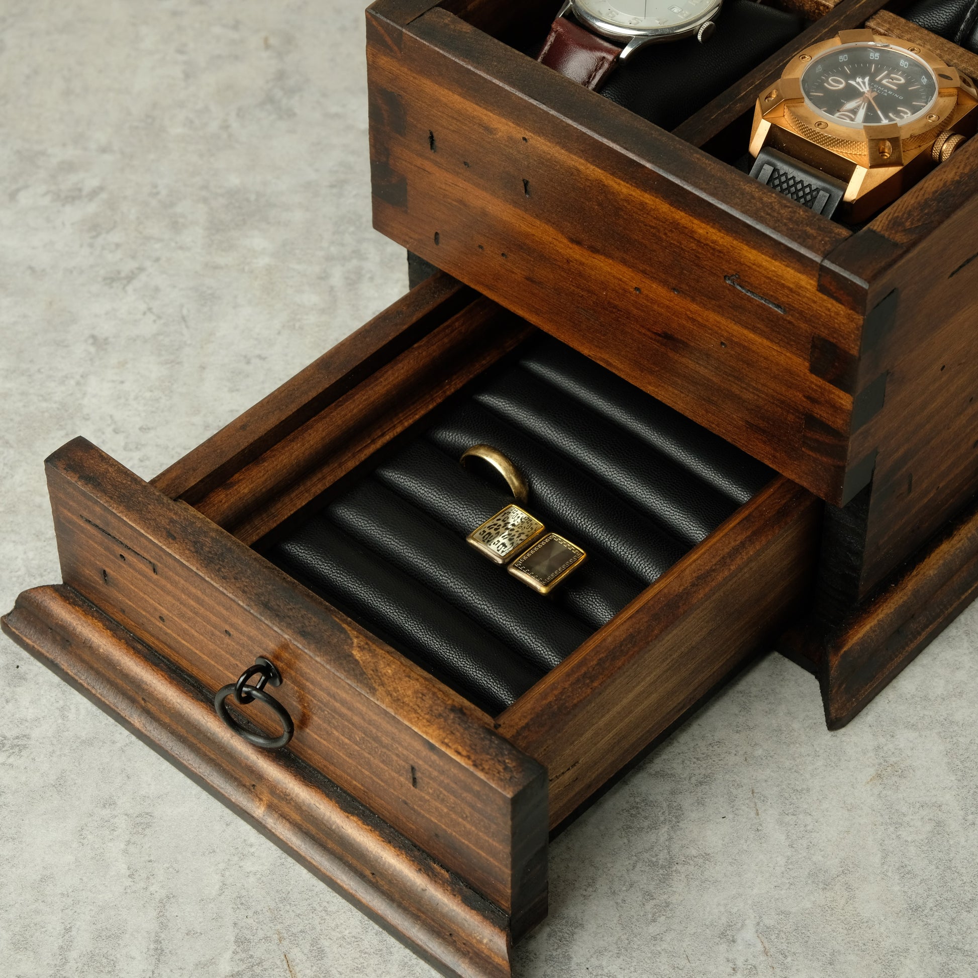 A rustic dark brown wooden watch and jewelry organizer with an open drawer showing a black ring roll holding a gold ring, with a watch and another watch partially visible in the upper compartments, set on a light gray concrete surface.