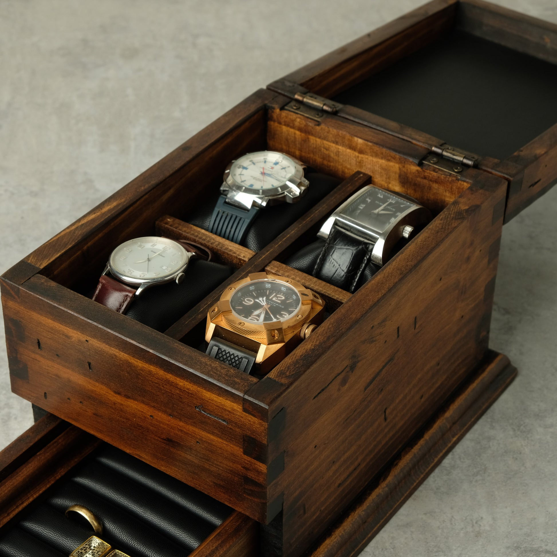 A dark brown wooden watch storage box with an open top tray holding several wristwatches and a lower drawer with black roll-up slots, photographed on a gray surface.
