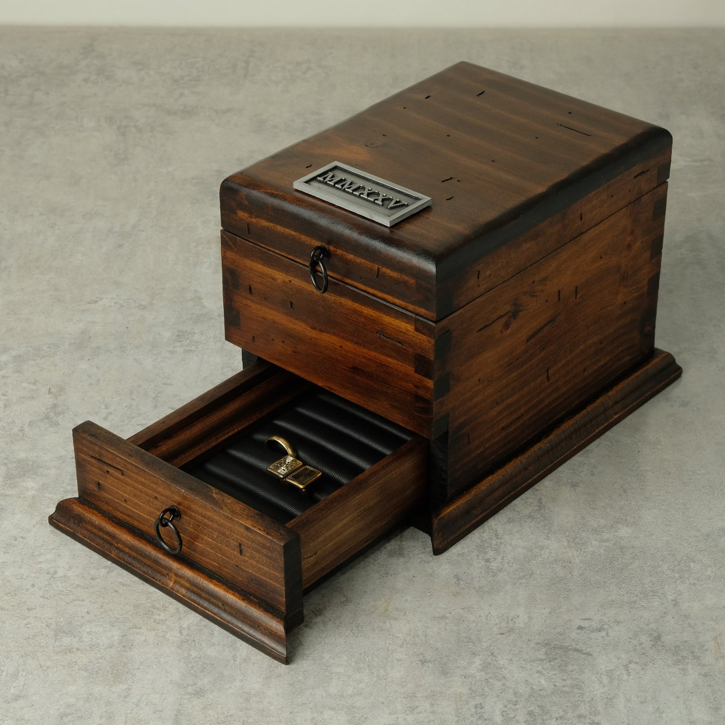 A dark brown wooden watch box with one drawer pulled open, showing a black ring roll inside. The box has a silver metal plaque that can be customized  on the top, and dark metal ring pulls on the drawer fronts, set against a neutral gray concrete background.