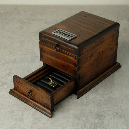 A dark brown wooden watch box with one drawer pulled open, showing a black ring roll inside. The box has a silver metal plaque that can be customized  on the top, and dark metal ring pulls on the drawer fronts, set against a neutral gray concrete background.