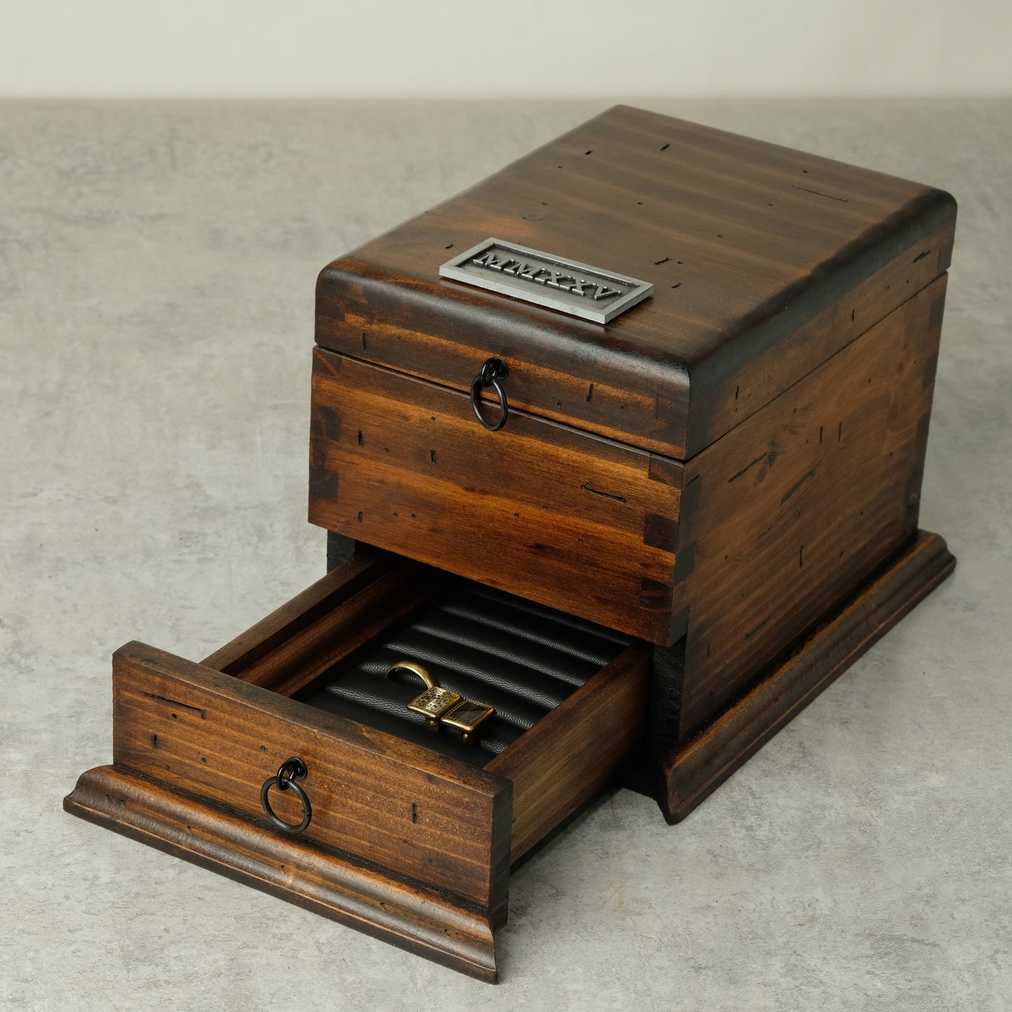 A reclaimed wooden watch box with a top drawer and an open bottom drawer showing a ring roll and padlock, photographed on a light gray textured surface.