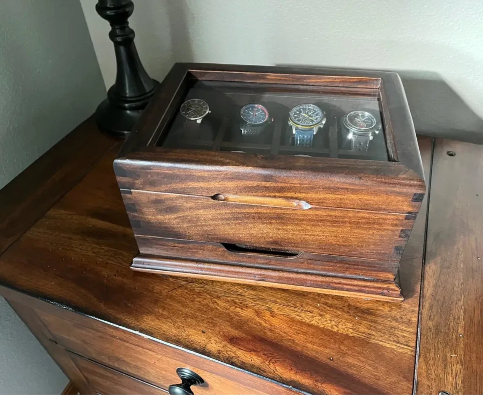 A compact handcrafted watch box displayed on a wooden dresser beside a lamp.