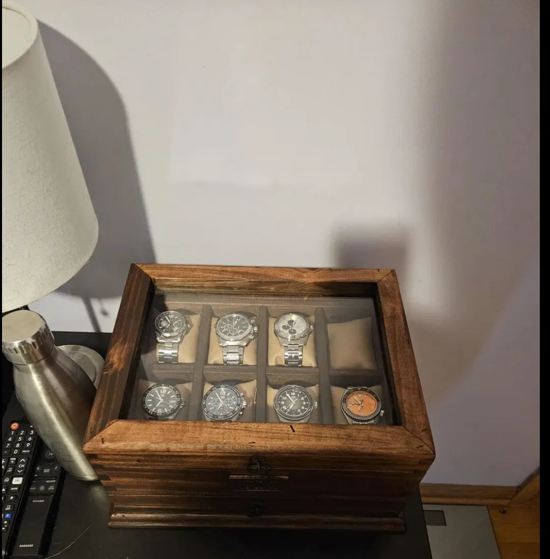A glass-lid watch box holding seven watches beside a lamp and desk accessories.